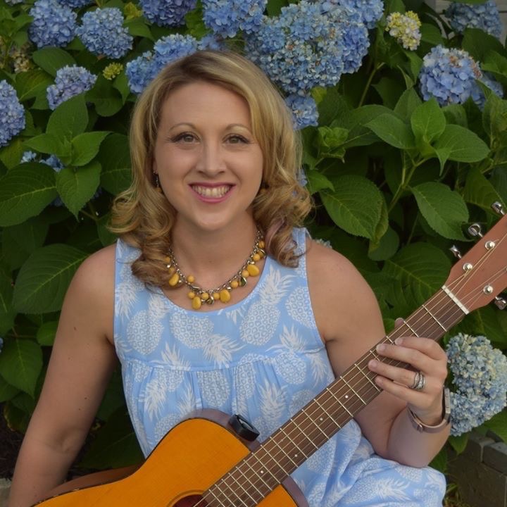 Laura Theismann with acoustic guitar in front of blue hydrangeas